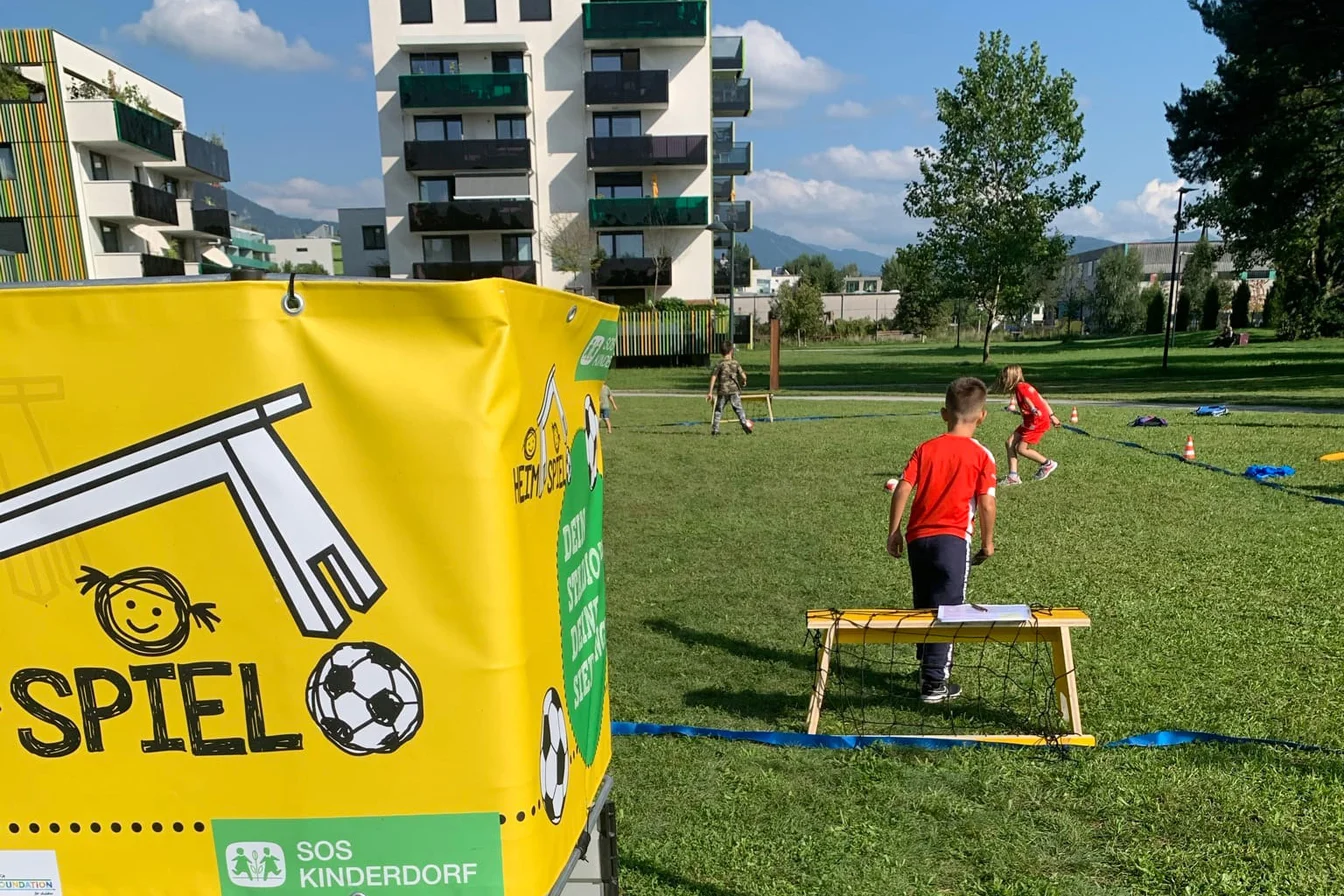 Kinder spielen Fußball auf einer Wiese in der Nähe von Wohnhäusern in Österreich.